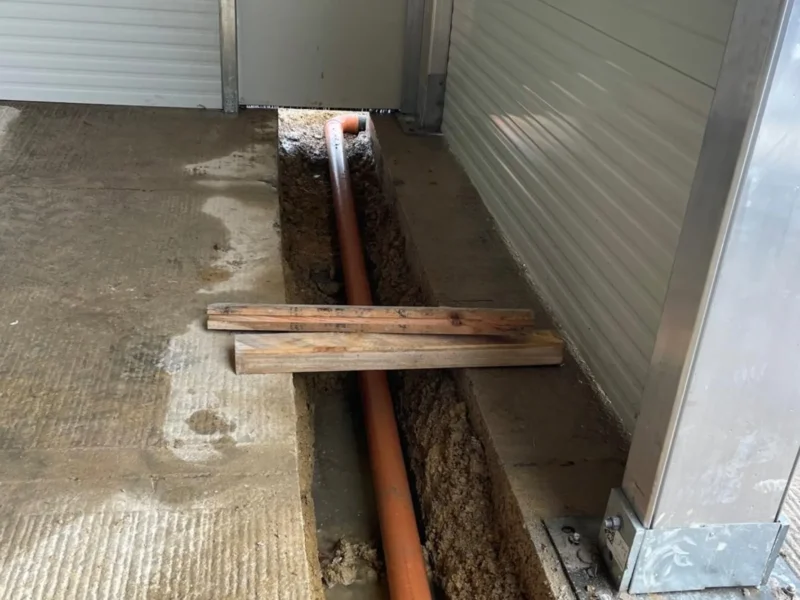 A section of orange underground pipe laid in a trench running along the base of a metal-clad building wall. Wooden planks rest across the trench, and the concrete floor shows signs of recent cutting and excavation.