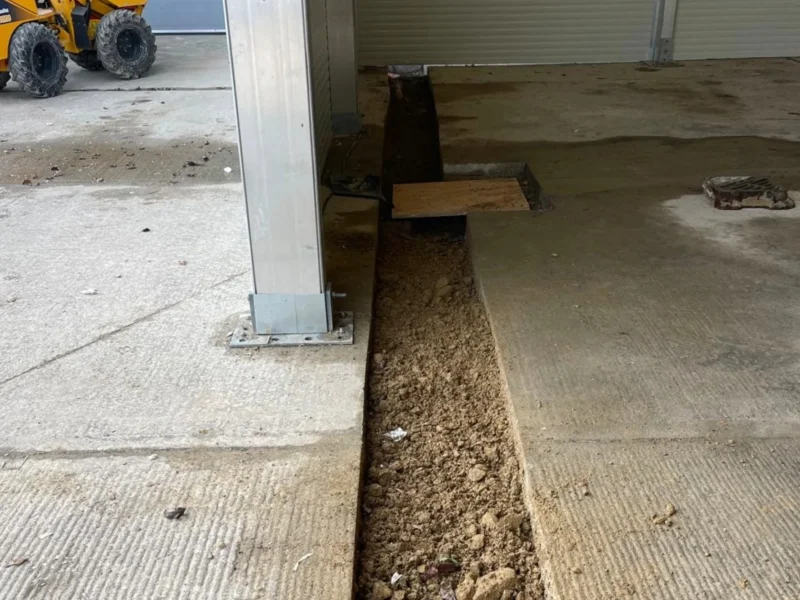 A trench cut into a concrete floor running alongside a metal support column. Soil is visible in the trench, and construction equipment can be seen in the background outside the building.