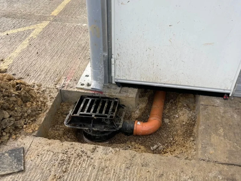 A close-up view of a drainage installation beside a building wall. An orange underground pipe connects to a black drain cover set into a concrete recess, with exposed soil and rubble around the opening.